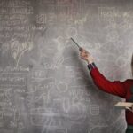 Quantum Physics - Serious female teacher wearing old fashioned dress and eyeglasses standing with book while pointing at chalkboard with schemes and looking at camera