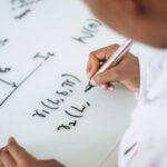 Quantum Physics - From above anonymous ethnic student wearing uniform and solving problem in chemistry while writing formula on white table in lab