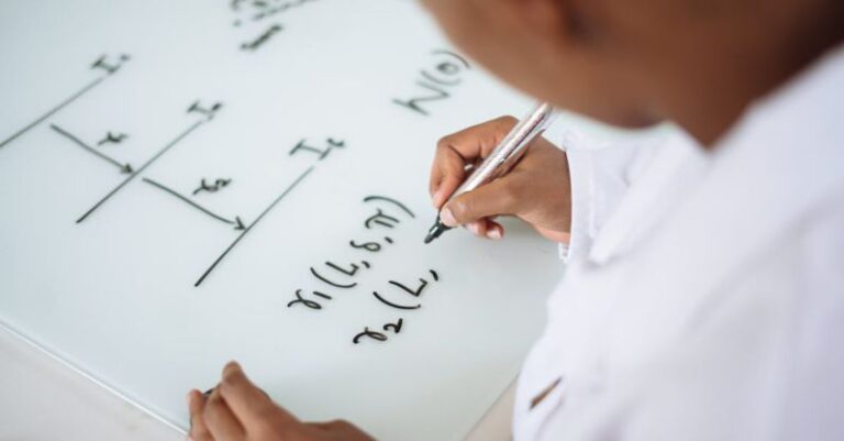 Quantum Physics - From above anonymous ethnic student wearing uniform and solving problem in chemistry while writing formula on white table in lab